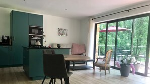 Una cocina moderna con armarios azul verdosos, un comedor con mesa y sillas de madera, y un amplio ventanal corredero que da a un patio exterior con plantas y un paraguas rojo.