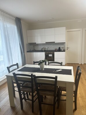 Uma área de refeições com mesa, cadeiras, um vaso de planta e uma cozinha ao fundo.