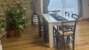 A dining area with a white table and dark wooden chairs, a stone wall, and a large window with curtains. - Podere Belsogno-apartment rose garden (Montalcino)
