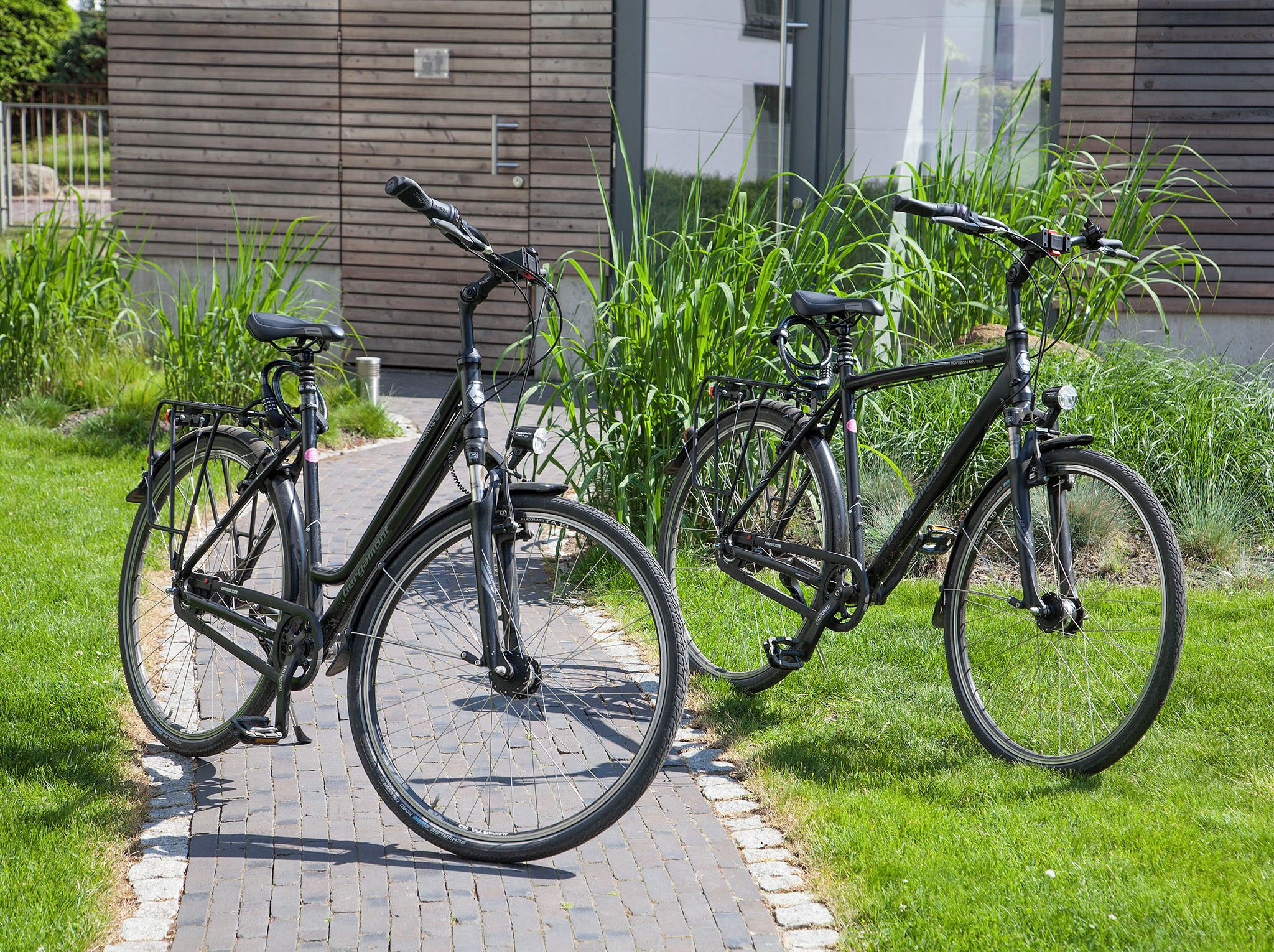 Two bicycles parked on a paved path with grass on either side, in front of a modern building with horizontal siding.