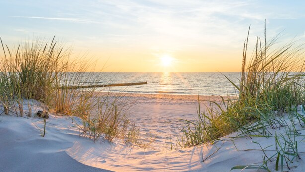 A beach scene with sand dunes and grasses, a wooden pier extending into the water, and the sun setting on the horizon. - Baltic Cloud, Villa Augustine (Binz)
