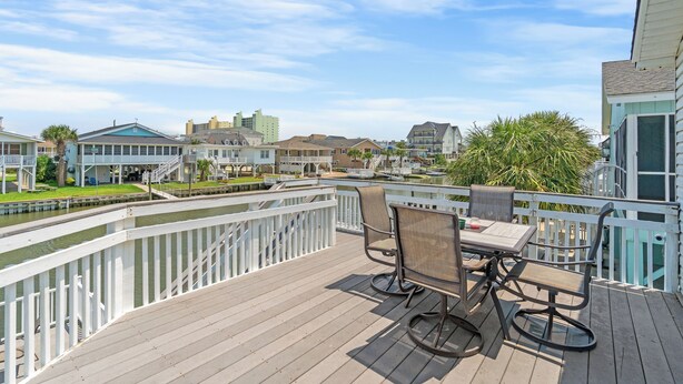 A wooden deck with a table and chairs, overlooking a marina with boats and buildings in the background. - Driftwood Too - 4 Bed/2 Bath Home on the Channel in Cherry Grove (North Myrtle Beach)