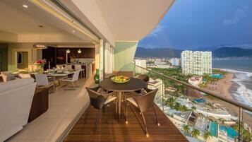 A balcony with a dining area, a kitchen, and a city view with a beach and buildings.