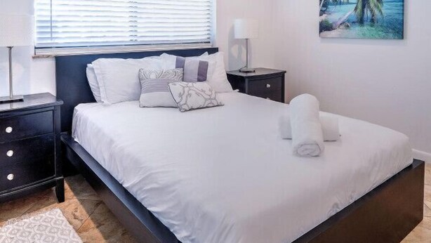 A bedroom with a bed, nightstands, a wall-mounted lamp, a ceiling fan, a window with blinds, and a painting of a beach scene.