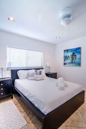 A bedroom with a bed, nightstands, a wall-mounted lamp, a ceiling fan, a window with blinds, and a painting of a beach scene. - A lg 1bed Off Las Olas on the water free kayaks (Fort Lauderdale)