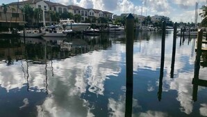 A marina with boats docked, a calm water surface reflecting the sky, and buildings in the background.