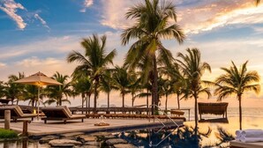 Zona junto a la piscina con sillones de descanso, palmeras y vista al océano.