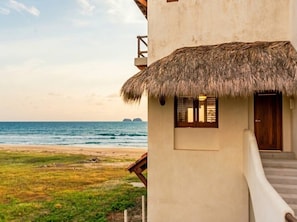 A beachfront property with a thatched-roof structure, a staircase leading to a wooden door, and a view of the ocean.