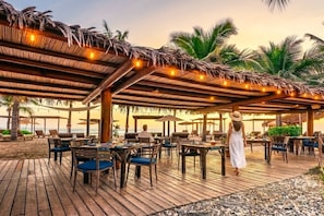 An outdoor dining area with wooden tables and chairs, a thatched roof, and palm trees.