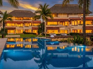 A resort with a swimming pool, palm trees, and buildings with thatched roofs.
