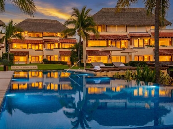 A resort with a swimming pool, palm trees, and buildings with thatched roofs.