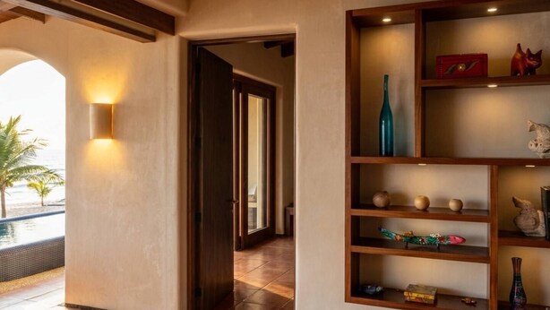 Un pasillo con una puerta de madera que da a una habitación, un estante con diversos objetos decorativos y vistas a una zona de piscina.