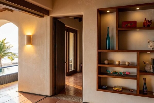 A hallway with a wooden door leading to a room, a shelf with various decorative items, and a view of a pool area.
