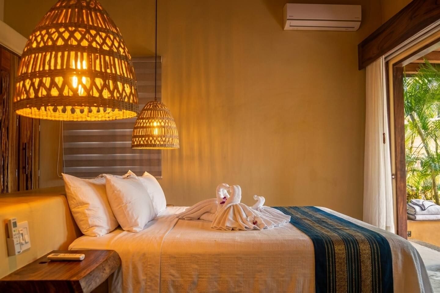 A hotel room with a bed, two hanging lamps, a window with a view, and an air conditioning unit.