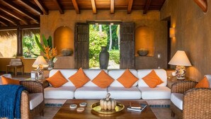 Ein großzügiger Wohnbereich mit einem weißen Sofa, das mit orangefarbenen Kissen geschmückt ist, einer Holzbalkendecke und Blick auf üppiges Grün draußen.