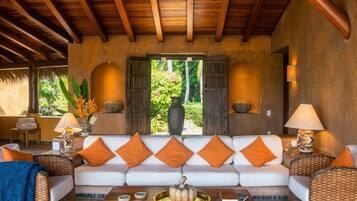 A spacious living area with a white sofa adorned with orange cushions, a wooden ceiling, and a view of lush greenery outside.