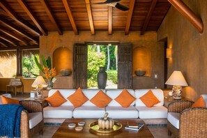 A spacious living area with a white sofa adorned with orange cushions, a wooden ceiling, and a view of lush greenery outside. - Casamar, beach front estate on Troncones Point (La Majahua)