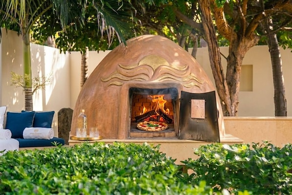 An outdoor pizza oven with a fire, surrounded by greenery and a sofa with cushions.