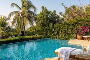 A clear blue pool with a lounge chair and a table, surrounded by lush greenery and palm trees.