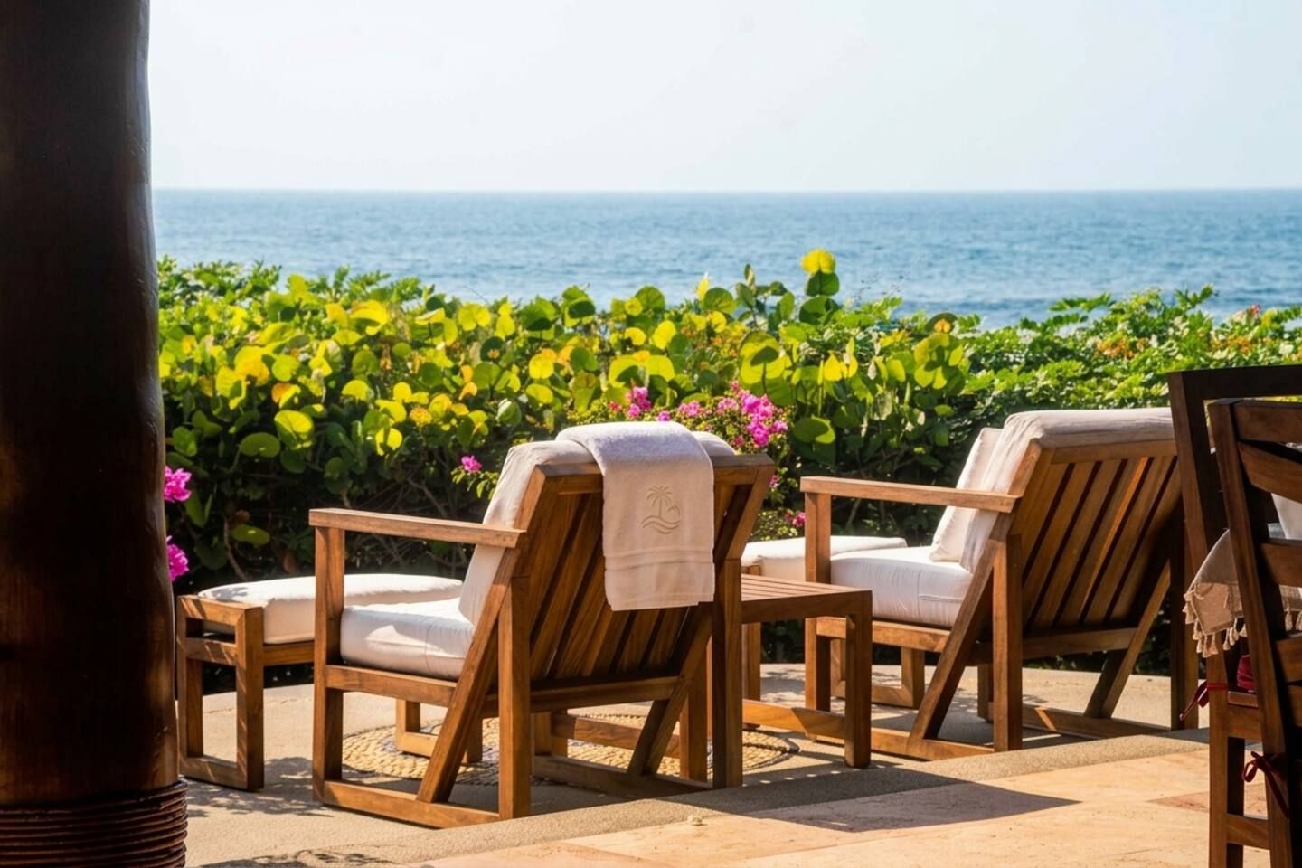 Two wooden lounge chairs with towels, positioned on a patio with a view of the sea and lush greenery.