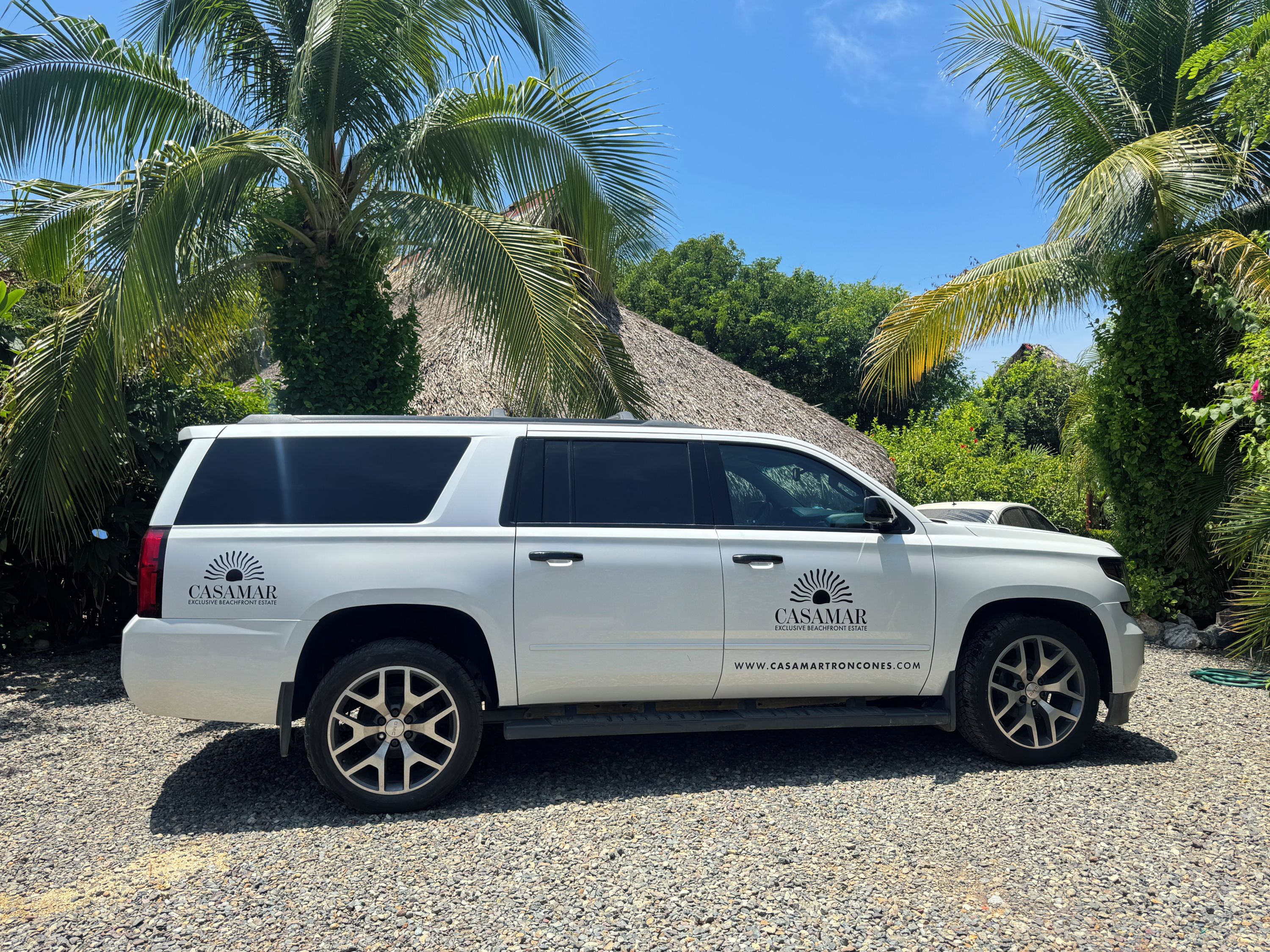 A white SUV parked on a gravel surface with palm trees and a clear blue sky in the background.