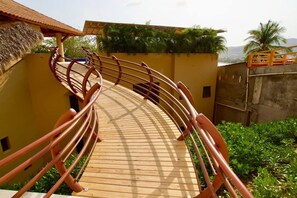 A wooden deck with metal railings, surrounded by greenery and a building with a thatched roof. - KauKan B7 Luxury Condo with Breathtaking View (Zihuatanejo)