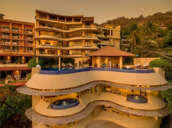 A multi-story hotel with a pool on the ground level, balconies on each floor, and a view of the surrounding landscape.