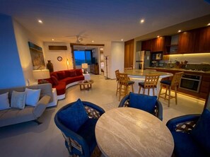 Una sala de estar moderna con un sofá, sillas y una mesa de comedor. Hay una cocina con armarios de madera y un refrigerador.