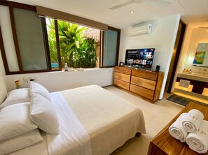 A bedroom with a bed, a TV, a wooden dresser, and a window with a view of greenery.
