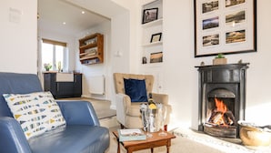 A living room with a blue sofa, a wooden coffee table with a book and glasses, a fireplace with a fire, and a kitchen area in the background. - Silva (Beaumaris)