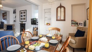 A dining area with a wooden table set for breakfast, surrounded by chairs. A fireplace is visible in the background, and a television is mounted on the wall. - Silva (Beaumaris)