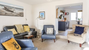 A living room with a sofa, armchairs, and a coffee table. There is a painting on the wall and a kitchen visible through an open door. - Silva (Beaumaris)