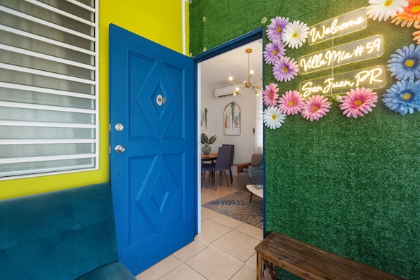 En blå dør med et neonlys der siger "Welcome Villa Mia #59 San Juan, PR" og en væg pyntet med kunstige blomster.
