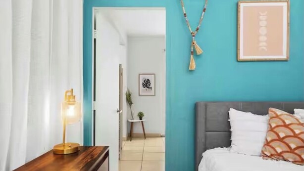 A bedroom with a bed, a bedside table with a lamp, a framed picture on the wall, and a door leading to another room. - Pool - Water tank -Solar panels  5 Min AiRpOrT & Isla Verde Beach! Watch Video - (Carolina)