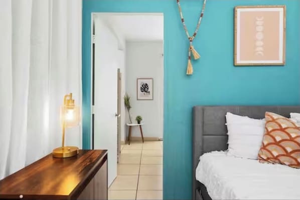 A bedroom with a bed, a bedside table with a lamp, a framed picture on the wall, and a door leading to another room. - Jump to San Juan AiRpOrT & Isla Verde Beach!Family sized -Watch Video - (Carolina)