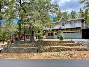 A two-story house with a garage, surrounded by trees and a stone retaining wall. - Bear Family Den • Game Room • Fenced • RV Parking (Big Bear)