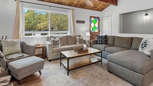 Ein gemütliches Wohnzimmer mit Holzbalkendecke, zwei Sofas, einem Couchtisch und einem Fenster mit Blick auf Bäume.