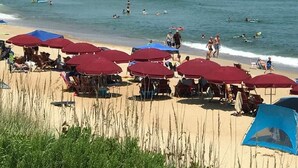 A beach with red umbrellas, a pier in the background, and people swimming and sunbathing. - BIS-Duck Resort - 2BR/2.5BA - Great Beach and Pools! (Duck)