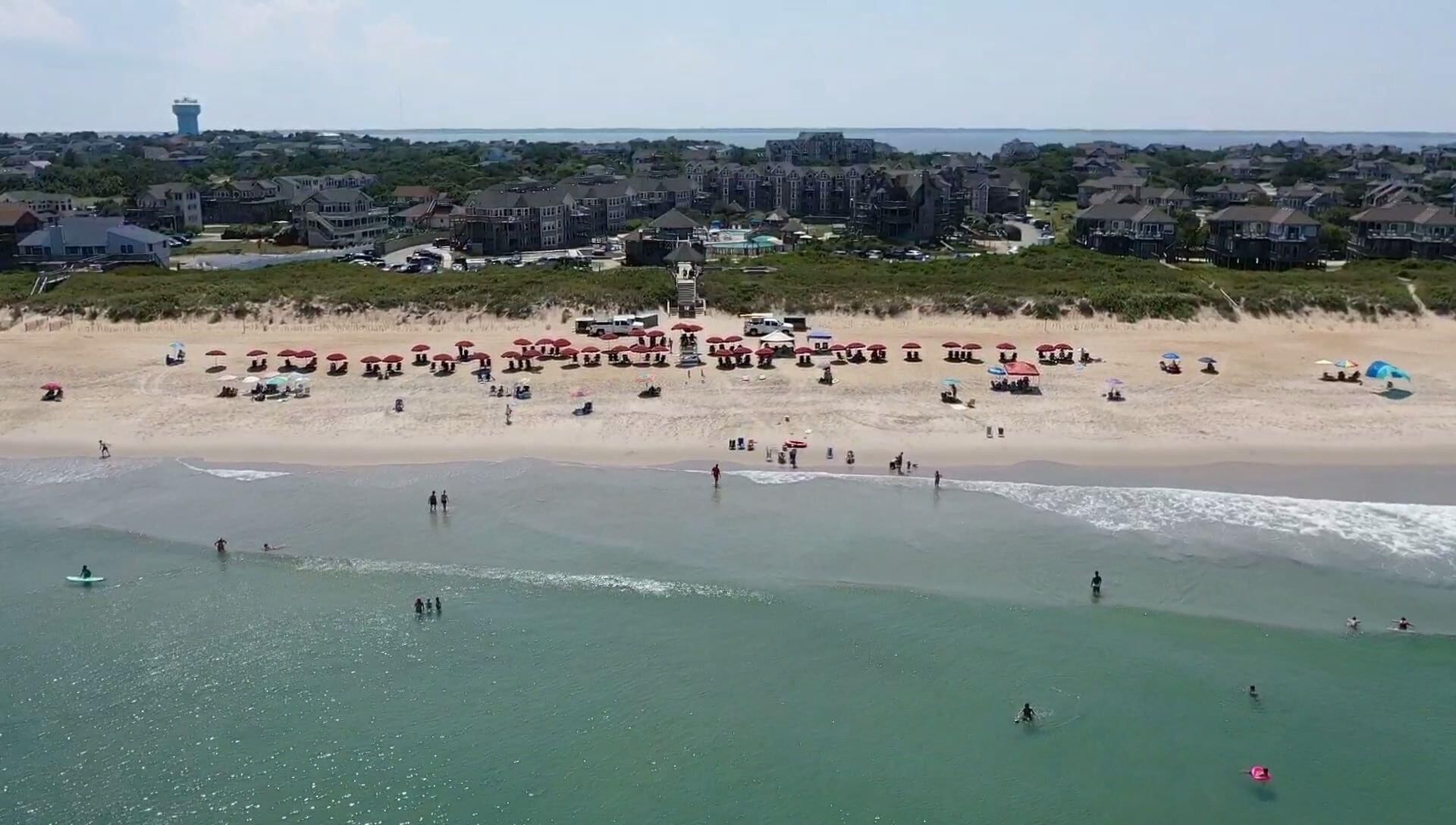 Ein Strand mit Sonnenschirmen und Menschen, ein Wasserturm im Hintergrund und Häuser in der Ferne.