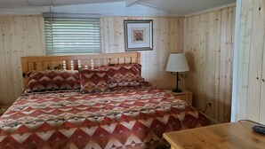 Een kamer met houten lambrisering, een bed, nachtkastje, lamp en een lijst met een afbeelding aan de muur.