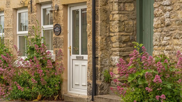 A stone building with a white door and windows, surrounded by pink flowering plants.