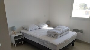 Una cama bien tendida con sábanas y almohadas blancas, una mesita de noche con lámpara, y un ventanal que deja entrar luz natural.
