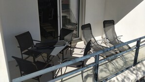 Un balcón con mesa y sillas, un ventanal corredera y barandilla de metal.