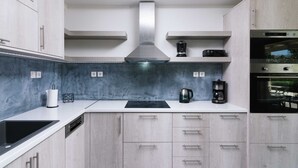 A modern kitchen with light wood cabinets, a stainless steel range hood, a built-in oven, and a coffee maker on the countertop. - Αmfithea Villas (Πετροκεφάλι)