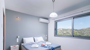 A bedroom with a bed, bedside tables, a window with a view of the countryside, and an air conditioning unit. - Αmfithea Villas (Πετροκεφάλι)