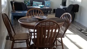 Un área de comedor con una mesa y sillas de madera, un sofá y un televisor en una habitación con piso de madera y ventanas grandes.