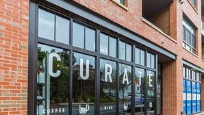 Un bâtiment doté de grandes fenêtres sur lesquelles est inscrit le mot « CURATE » et où figurent des panneaux indiquant « CAFÉ » et « VIN ».