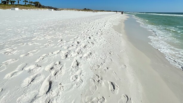Ein weiter Sandstrand mit Fußspuren, die zum Wasser führen, Palmen und einem strahlend blauen Himmel.
