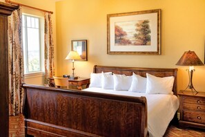 A hotel room with a wooden bed, a desk with a lamp, a bedside table with a lamp, and a framed painting on the wall.