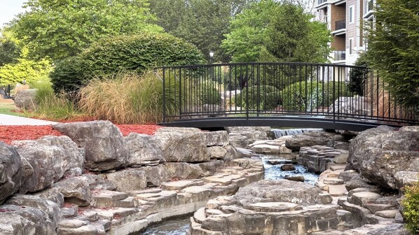 Un espace paysager agrémenté d’une cascade en pierre, d’un pont métallique et d’une bordure de paillis rouge.